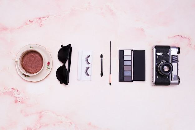 Xícara de café; oculos escuros; cílios; pincel de maquiagem; paleta da sombra e câmera vintage no pano de fundo texturizado rosa