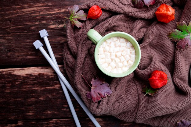 Xícara de café ou chocolate quente com marshmallow perto do cobertor de malha e agulhas.