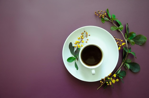 Xícara de café plana e em um pires redondo encontra-se um galho com pequenas flores e folhas amarelas