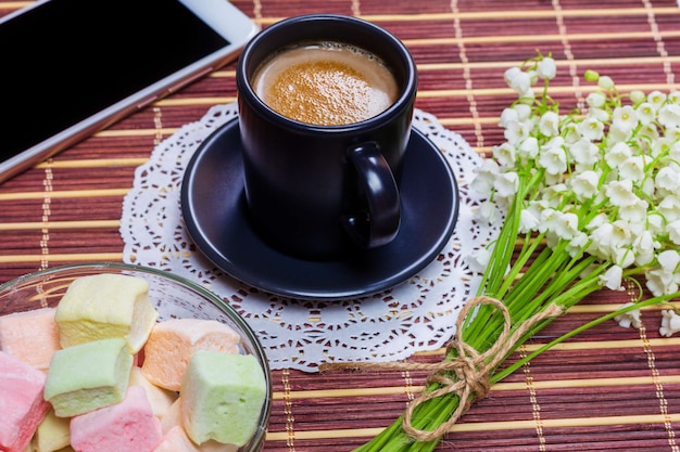 Xícara de café preta com leite em um pires, marshmallow, um buquê de flores de lírio do vale e smartphone em cima da mesa