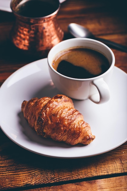 Xícara de café preto e croissant fresco na mesa de madeira