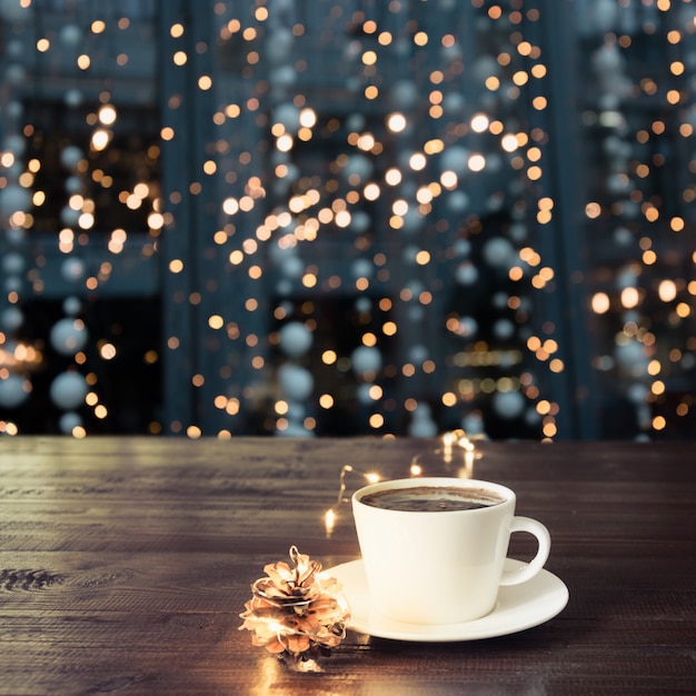 Xícara de café preto na mesa de madeira no café. Luzes de Natal e guirlanda de ouro sobre fundo.