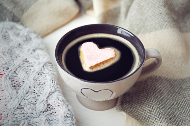 Xícara de café quente com marshmallow de coração e cachecóis quentes sobre fundo claro de madeira close-up