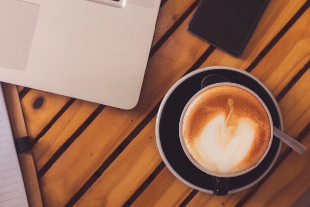 Xícara de café quente de vista superior com smarthphone de laptop borrado e caderno na mesa de madeira