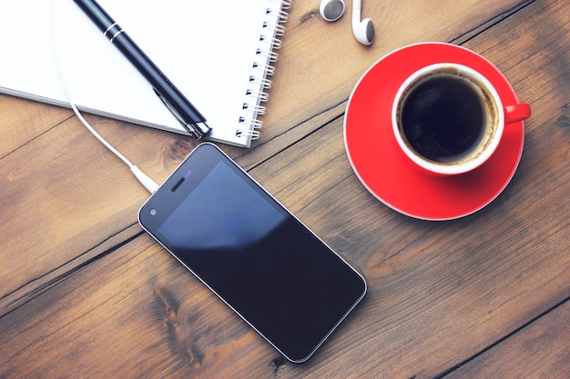 Xícara de café, telefone, fone de ouvido, caderno e caneta na mesa de madeira