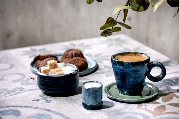 Xícara de café turco preto com leite, cubos de açúcar e biscoitos na mesa de cerâmica ornamentada com galhos de eucalipto. luz solar, sombras