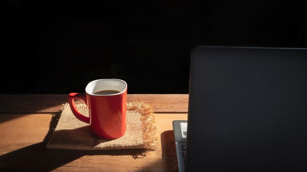 Xícara de café vermelha e laptop preto desfocado na mesa de madeira com luz solar e sombra em fundo preto