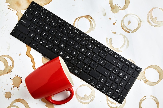 Xícara de café vermelha virada em um teclado de computador em uma mesa com manchas de café