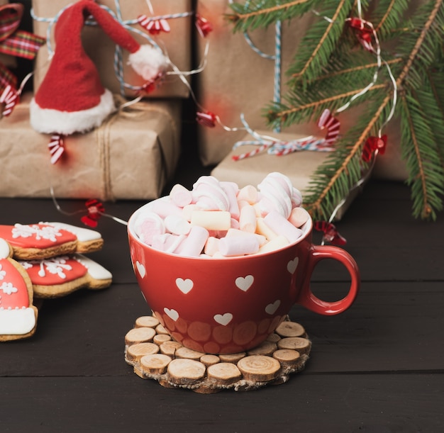 Xícara de cerâmica vermelha com cacau e marshmallows, atrás de uma caixa de presente e um brinquedo de Natal