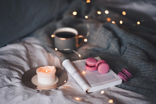 Xícara de chá de blac com vela acesa perfumada com livro de papel aberto e macaroons de fruta rosa na cama na malha jumper sobre luzes de brilho no fundo close-up. bom dia. atmosfera caseira romântica aconchegante.