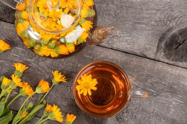 Xícara de chá de ervas, bule transparente e flores de calêndula em fundo de madeira.