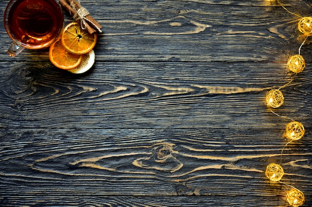 Xícara de chá, fatias secas de laranja e limão e paus de canela com luzes de Natal em um fundo escuro de madeira. Fundo festivo. Vista do topo.