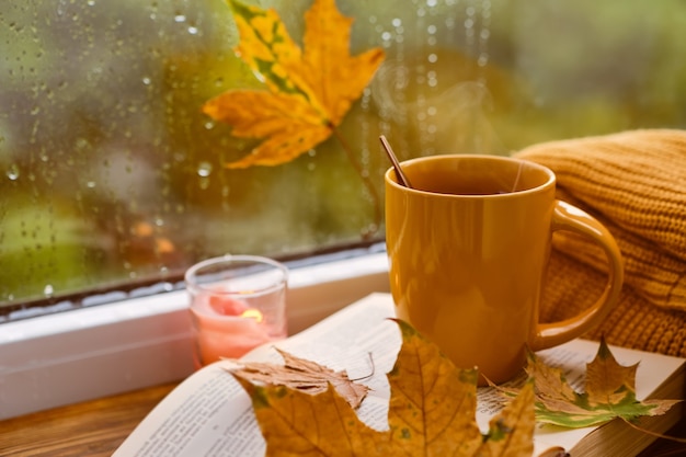 Xícara de chá, livro, folhas de outono e vela no peitoril da janela em casa