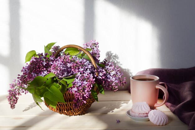 Xícara de chá ou café com marshmallows rosa doces e buquê de lilás em uma cesta. Café da manhã.