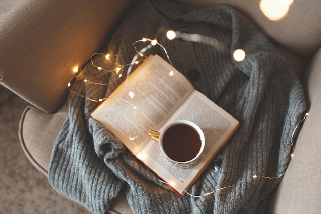 Xícara de chá preto fresco permanecer no livro de papel aberto com a camisola de lã tricotada em uma cadeira aconchegante sobre brilho closeup de luzes de Natal. Vista do topo. Café da manhã de manhã cedo. Atmosfera caseira confortável de inverno.
