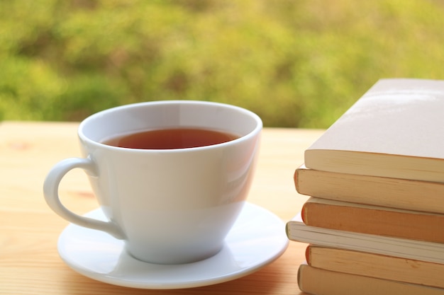 Xícara de chá quente com pilha de livros na mesa de madeira com fundo desfocado de plantas no jardim