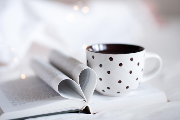 Xícara de chá quente fica no livro de papel aberto com páginas dobradas em forma de coração sobre luzes brilhantes na cama close-up ambiente aconchegante em casa