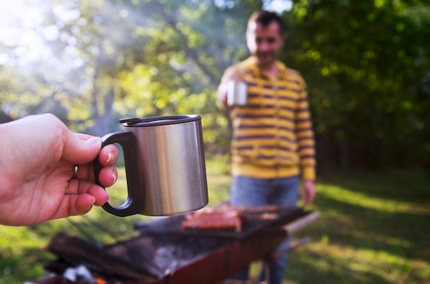 Xícara de metal de piquenique na mão de uma mulher torcendo com amigo masculino na fumaça de churrasco