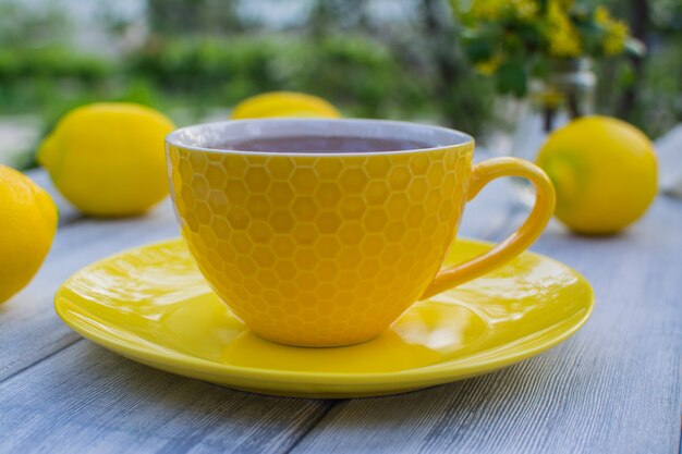 Xícara de porcelana amarela com chá com um pires amarelo em uma mesa de madeira No fundo estão limões amarelos