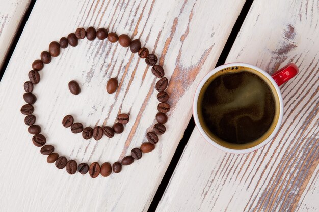 Xícara de vista superior de café quente e rosto sorridente. Superfície de madeira branca.