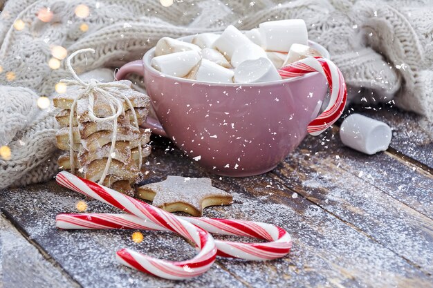 Foto xícara grande de cappuccino com marshmallow, bastões de doces e biscoitos na mesa de madeira marrom com luzes de natal.