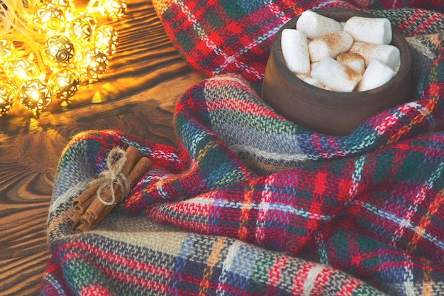 Xícara grande de chocolate quente com marshmallow, canela e cobertor quente em um vintage antigo de madeira e luz de natal. arranjo acolhedor de natal ou outono.