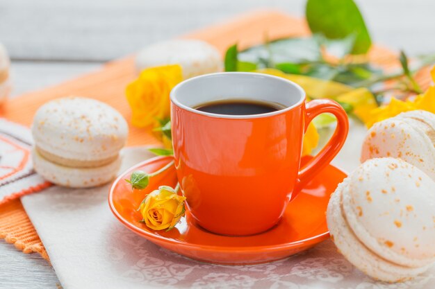 Xícara laranja de café preto, flores rosas amarelas e macaroons franceses pastel doces na mesa de madeira branca