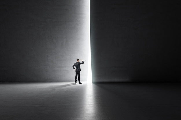 Éxito empresarial y mirar hacia el concepto futuro con un hombre de negocios mirando a través de un agujero de luz en una pared oscura en un espacioso salón vacío