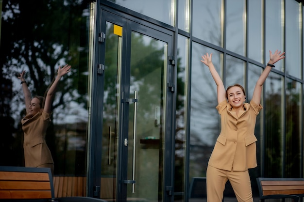 Éxito empresarial - Mujer de negocios joven feliz celebrando los logros de su carrera laboral con ambas manos contra el edificio de oficinas.