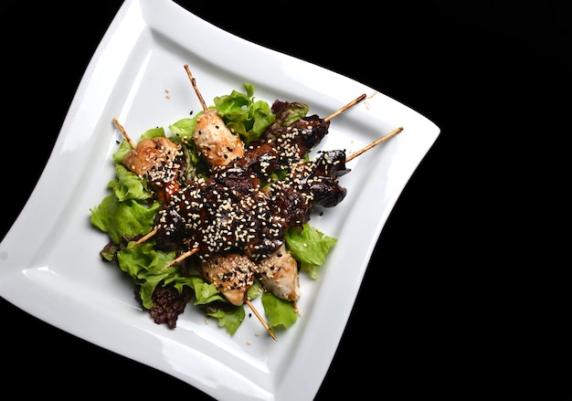 Yakitori con semillas de sésamo en un plato. Carne de pollo frito e hígado de pollo en primer plano de palos aislado sobre un fondo negro. Vista superior.