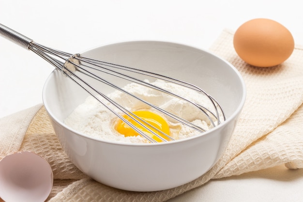Yema de huevo con harina y batir en un bol. Cáscara de huevo y huevo en la mesa. Fondo blanco. De cerca
