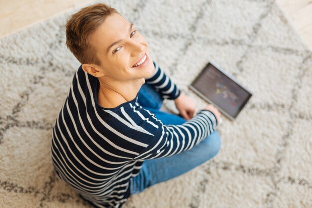 Yo estoy feliz. Exuberante rubio bien construido elegante joven sonriendo y sentado en el suelo y usando su tableta