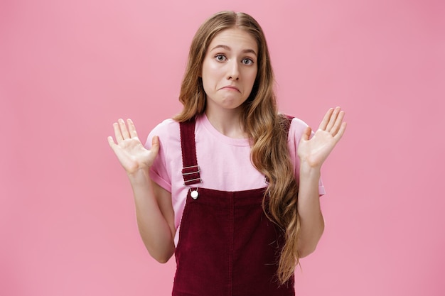 Yo no estoy involucrado. Retrato de mujer linda inocente inconsciente y desorientada que levanta las palmas en señal de rendición tirando de los labios hacia abajo como si estuviera confundido e inseguro de lo que sucede sin tener idea sobre la pared rosada.