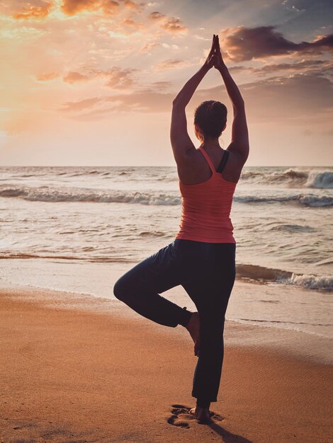 Yoga al aire libre mujer haciendo Hatha yoga asana Vrikshasana árbol postura en la playa tropical al atardecer