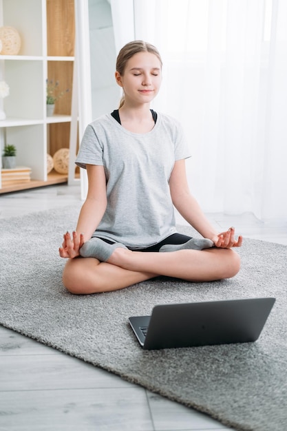 Yoga en casa para niños Meditación en línea Tutorial de capacitación virtual Niña tranquila y relajada sentada con las piernas cruzadas en loto en el piso con una computadora portátil en el interior de una habitación luminosa