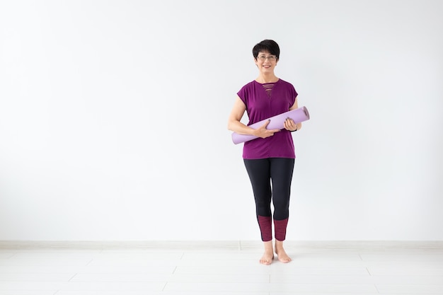 Yoga, concepto de peopel - mujer de mediana edad sosteniendo estera después de una clase de yoga en una superficie blanca con espacio de copia