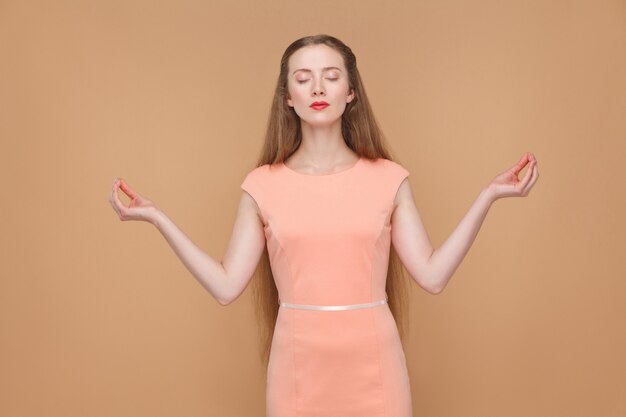 Yoga, Entspannung und Meditation. Porträt einer emotionalen süßen, schönen Frau mit Make-up und langen Haaren im rosa Kleid. Indoor, Studioaufnahme, isoliert auf hellbraunem oder beigem Hintergrund.