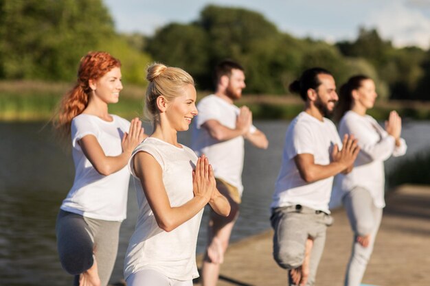 Yoga-, Fitness-, Sport- und gesundes Lebensstilkonzept – eine Gruppe von Menschen in einem Baum posiert auf einer Matte im Freien am Fluss- oder Seeliegeplatz