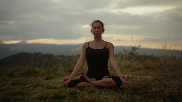 Yoga-Frau mit geschlossenen Augen meditiert im Freien. Fitte Frau macht Namaste-Yoga-Pose. Sportliches Mädchen sitzt in Lotus-Pose auf Gras. Entspannter Trainer praktiziert Yoga in den Bergen