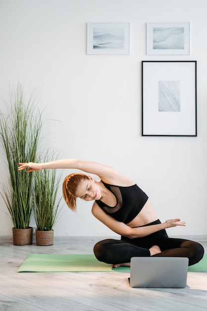 Yoga en línea Entrenamiento en el hogar Bienestar de la salud Mujer atlética en ropa deportiva viendo un tutorial web en una computadora portátil sentada con las piernas cruzadas en el suelo haciendo ejercicio de estiramiento en el interior de la luz