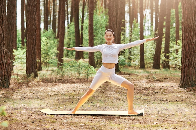 Yoga por la mañana al aire libre en el parque, instructor de joven hermosa mujer vestida con ropa deportiva elegante blanca de pie sobre la estera rodeada de árboles, deporte al aire libre.