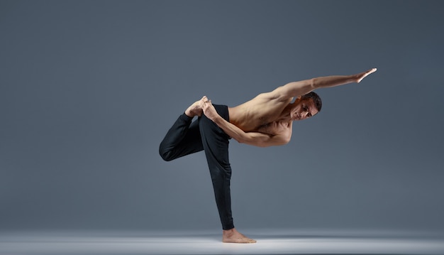 El yoga masculino mantiene el equilibrio en una pose difícil sobre una pierna, fondo gris. Hombre fuerte haciendo ejercicio yogui, entrenamiento de asanas, máxima concentración, estilo de vida saludable