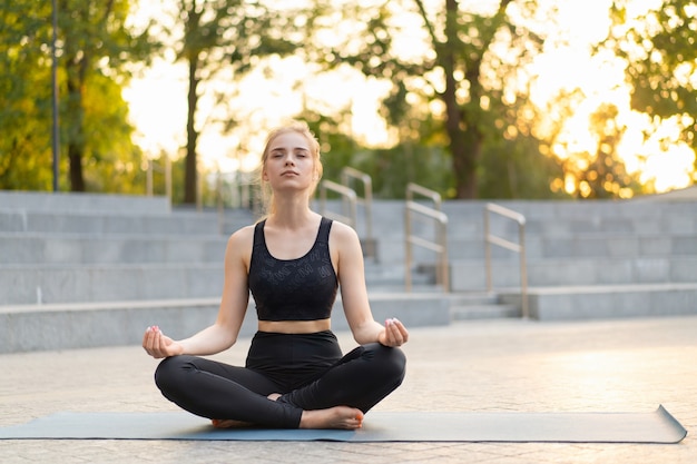 Yoga y meditación en la ciudad moderna Mujer caucásica Relajarse Posición de loto Sentado Estera de yoga al aire libre Parque de verano en piso de concreto Adulto joven Mujer en forma relajante después de la práctica de yoga Entrenamiento Siddhasana