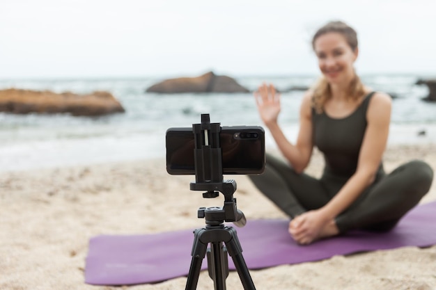 Yogalehrerin der jungen Frau leitet Online-Unterricht am Strand Sportfrau sendet zur Kamera des Smartphones, das auf einem Stativ steht Blogging vlog Gesunder Lebensstil