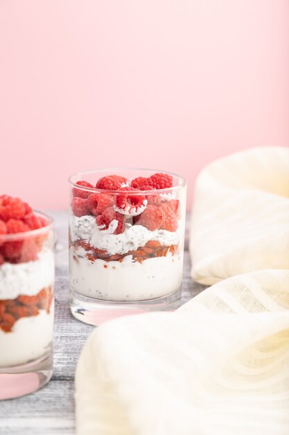 Yogur con frambuesa, bayas de goji y semillas de chía en vidrio sobre superficie gris y rosa y textil de lino