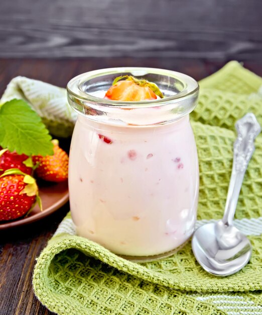 Yogur con fresas en un frasco de vidrio, cuchara sobre una servilleta, fresa y menta y un plato sobre un fondo de tablas de madera