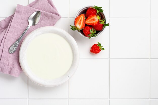 Yogur griego en un tazón blanco con ingredientes para hacer granola de desayuno y fresas frescas en el cuadro blanco. Vista superior.