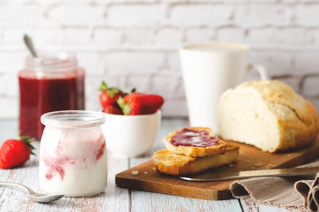 Yogur natural, fresas, mermelada de fresa, pan y una taza de café, sobre un fondo de madera.