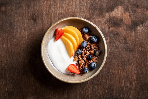 Yogur natural fresco con granola casera, melocotón, fresa y arándano en un bol sobre un fondo de madera oscura. El concepto de un desayuno saludable y nutritivo. Comida dietetica. Vista superior y espacio de copia.