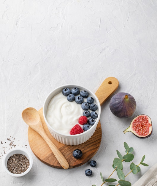 Yogurt fermentado súper sano natural con arándanos, higos, semillas de chía y frambuesa en un tazón blanco sobre una mesa gris claro. La imagen es espacio de copia y vista superior